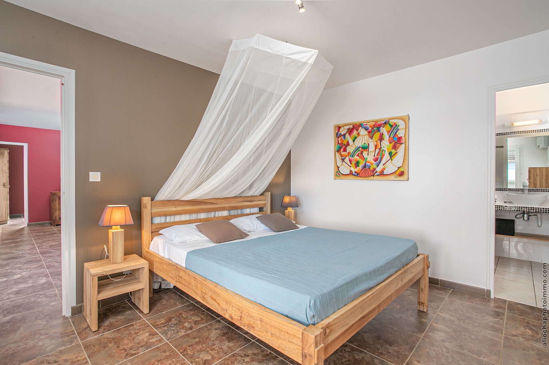 Une chambre à coucher avec un lit en bois, une table de chevet avec une lampe, un tableau accroché au mur, et une salle de bain visible à travers une porte ouverte.