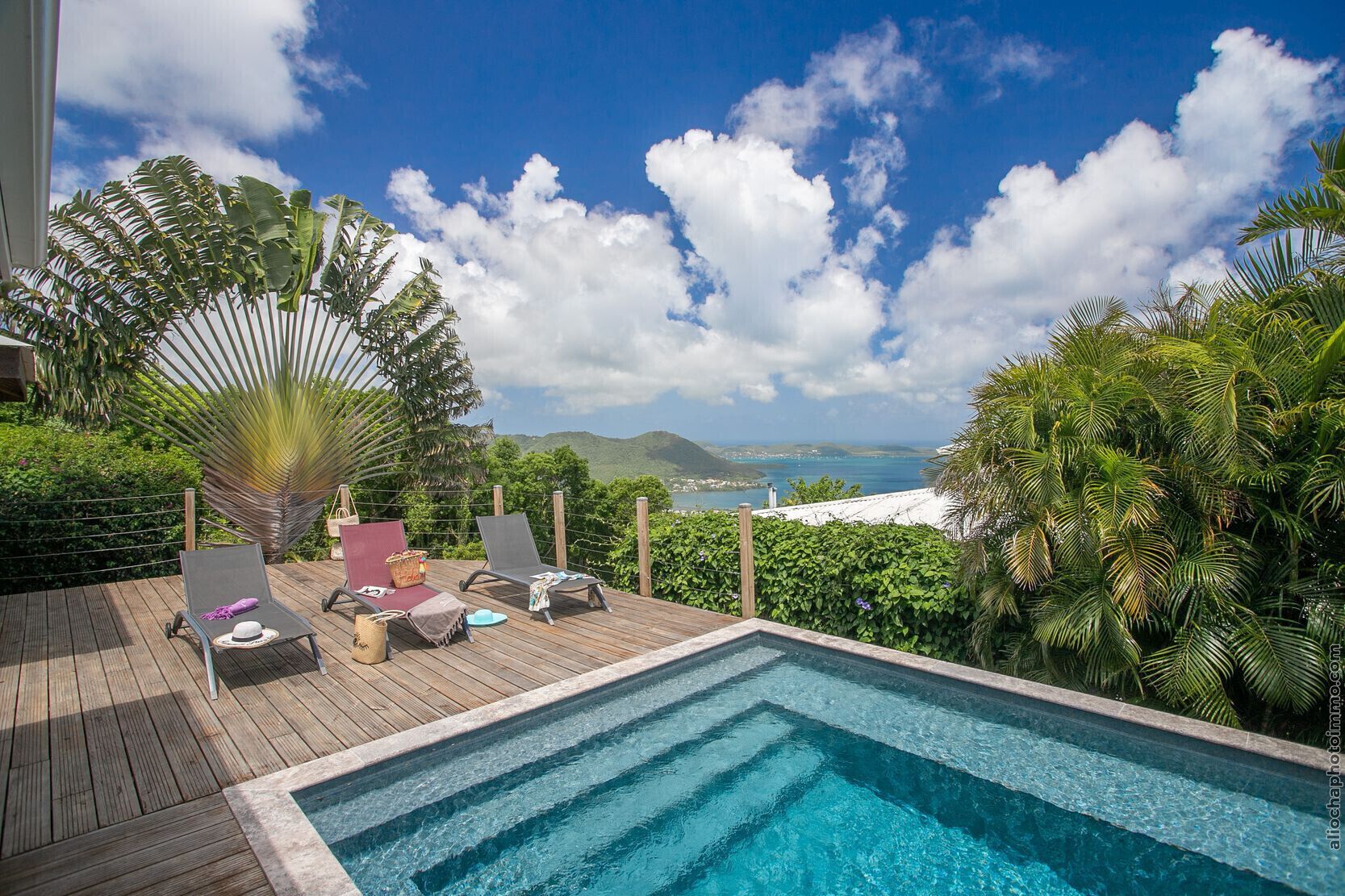 Un espace aménagé au bord de la piscine, avec des chaises longues, une terrasse en bois et une vue sur la mer et les collines.