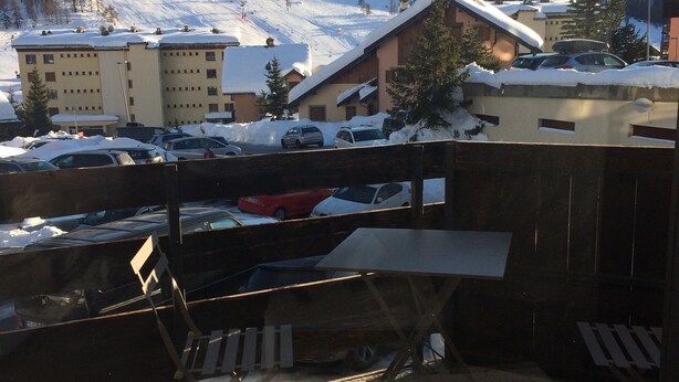 Uitzicht vanaf een balkon op een besneeuwd berglandschap, met gebouwen en geparkeerde auto's op de voorgrond.