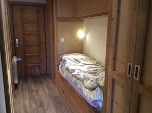 Une petite pièce étroite avec une porte en bois, un lit simple recouvert d’une courtepointe colorée et un luminaire placé au-dessus du lit.
