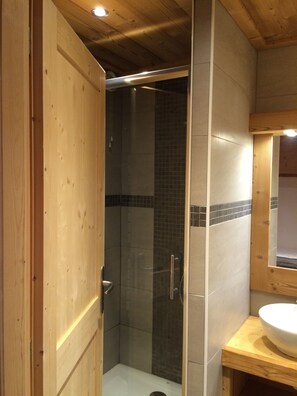 Une porte en bois donne accès à un espace douche, qui est délimité par une porte en verre et dont les murs sont recouverts de carreaux.