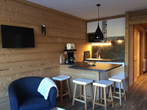 Une cuisine chaleureuse avec des murs en bois, un téléviseur à écran plat, une machine à café et un four à micro-ondes. On y trouve trois tabourets et une chaise bleue.