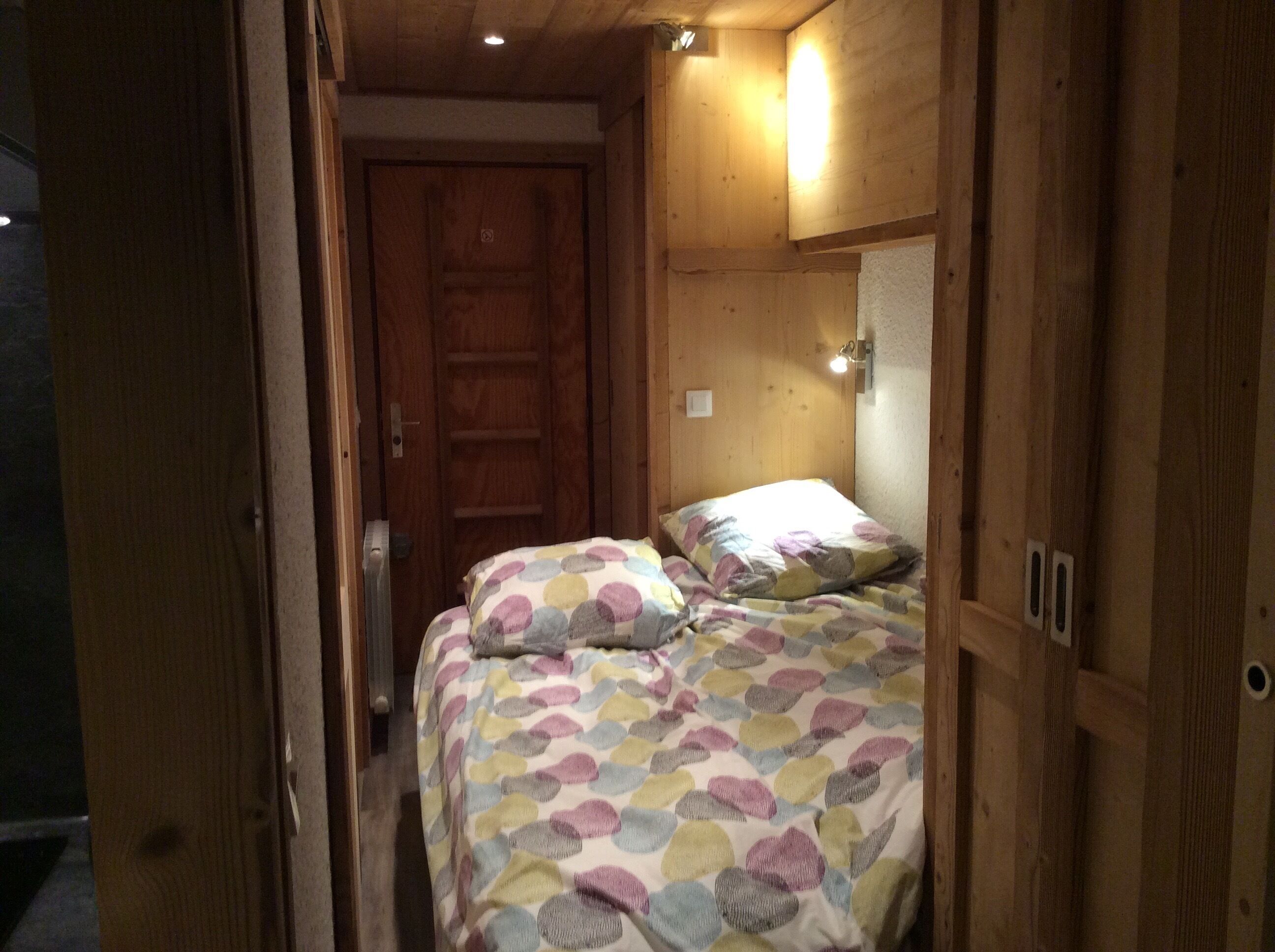 Una habitación pequeña y acogedora con una puerta de madera, una cama con una manta colorida y un estante con una luz.