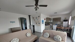 Ein modernes Wohnzimmer mit zwei beigefarbenen Sofas, einem Holztisch und einem Deckenventilator. Im Hintergrund ist eine Küche mit weißen Geräten zu sehen.