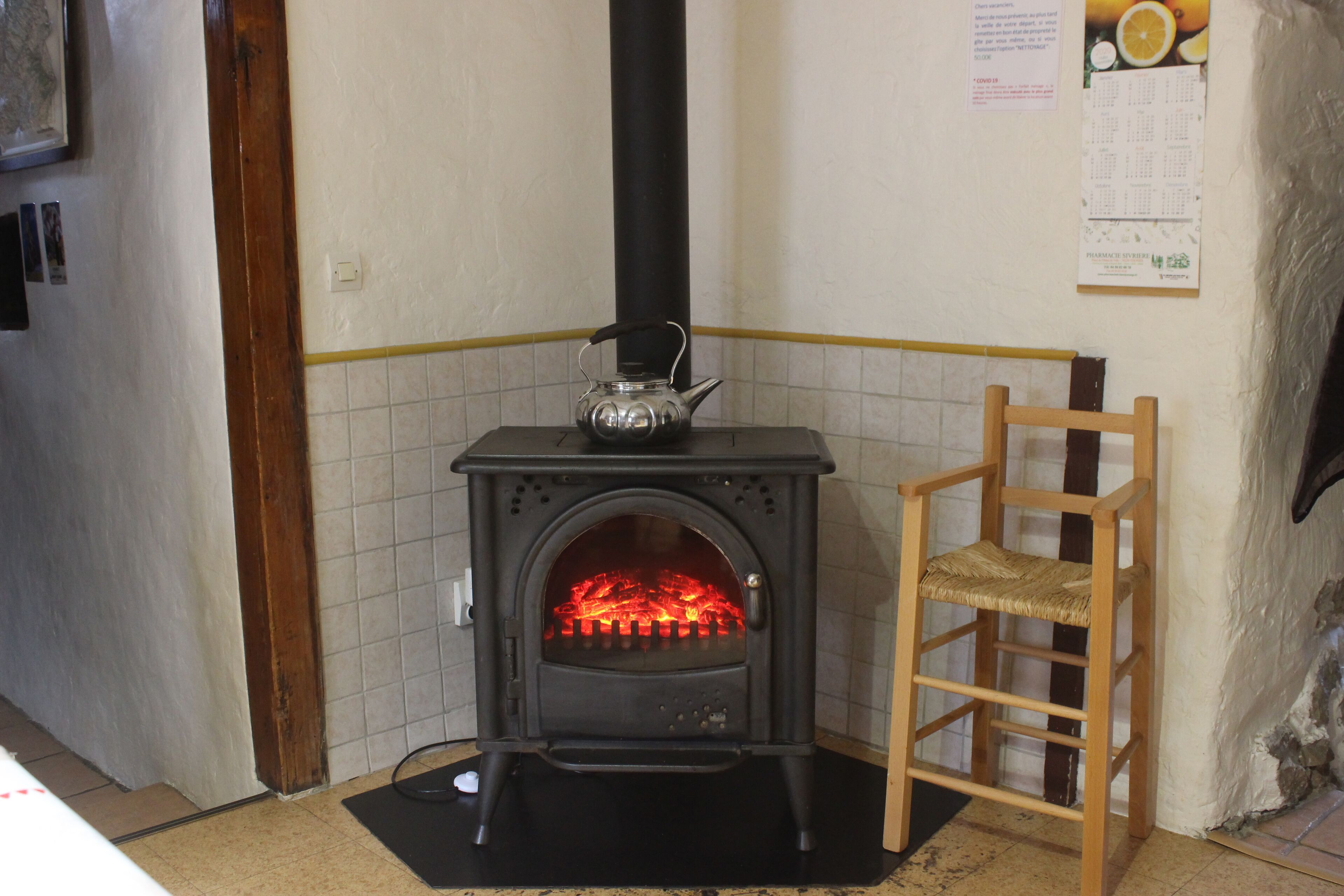 A wood-burning stove with a chimney, a wooden chair, and a tiled wall with a calendar.