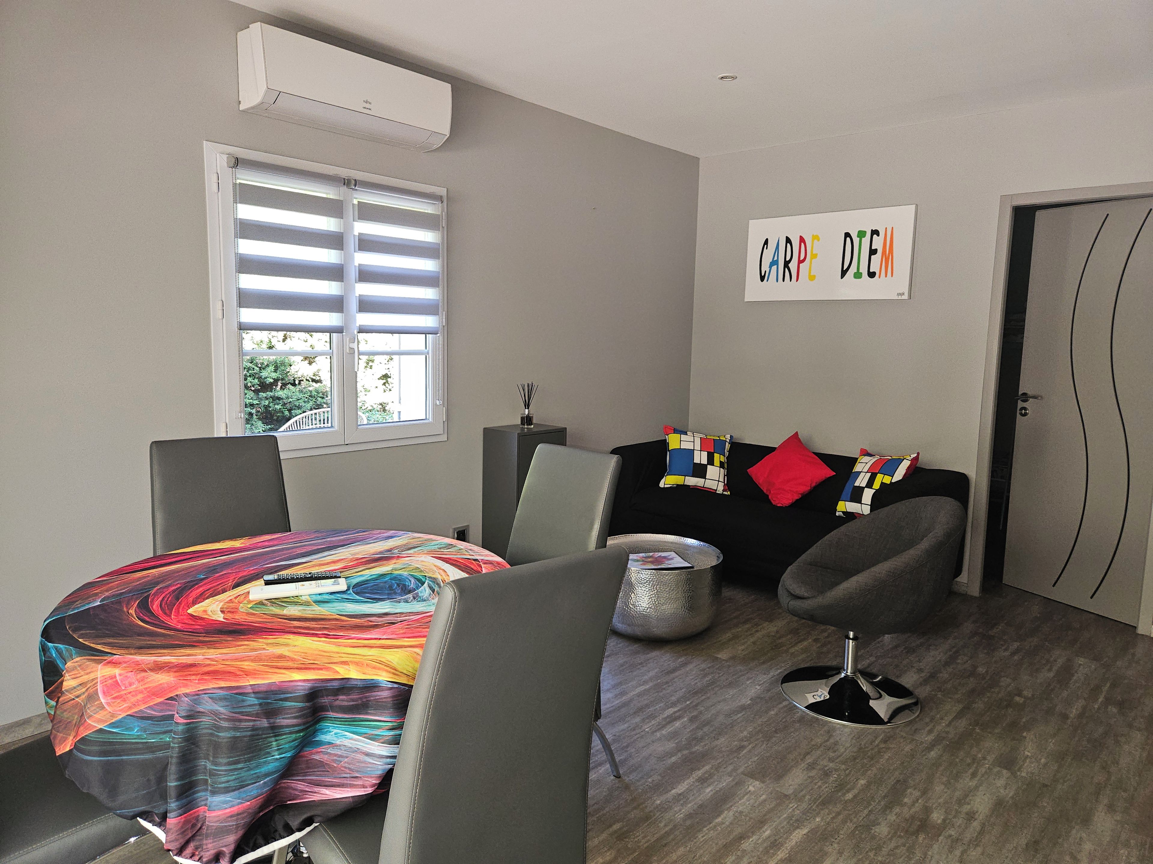 Ein modernes Wohnzimmer mit Esstisch, einer schwarzen Couch mit bunten Kissen, einem grauen Sessel und einer wandmontierten Klimaanlage.