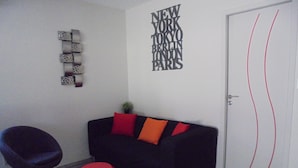 Un salon moderne avec un canapé noir, des coussins rouges et orange, un tabouret rouge et un mur orné de noms de villes.