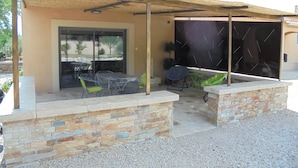 Un espace extérieur aménagé en patio, doté d’un coin salon abrité, d’un mur en pierre et d’une porte vitrée donnant sur l’intérieur.