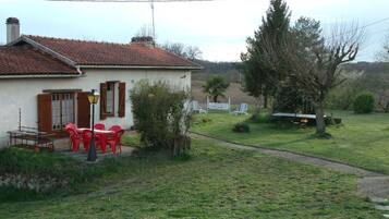 Una casa con techo de tejas rojas, un pequeño jardín con césped y algunos árboles, y un patio con sillas y una mesa rojos.