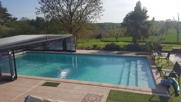 En swimmingpool med et glasomkranset område, omgivet af en terrasse med udendørs møbler og udsigt over et grønt landskab.