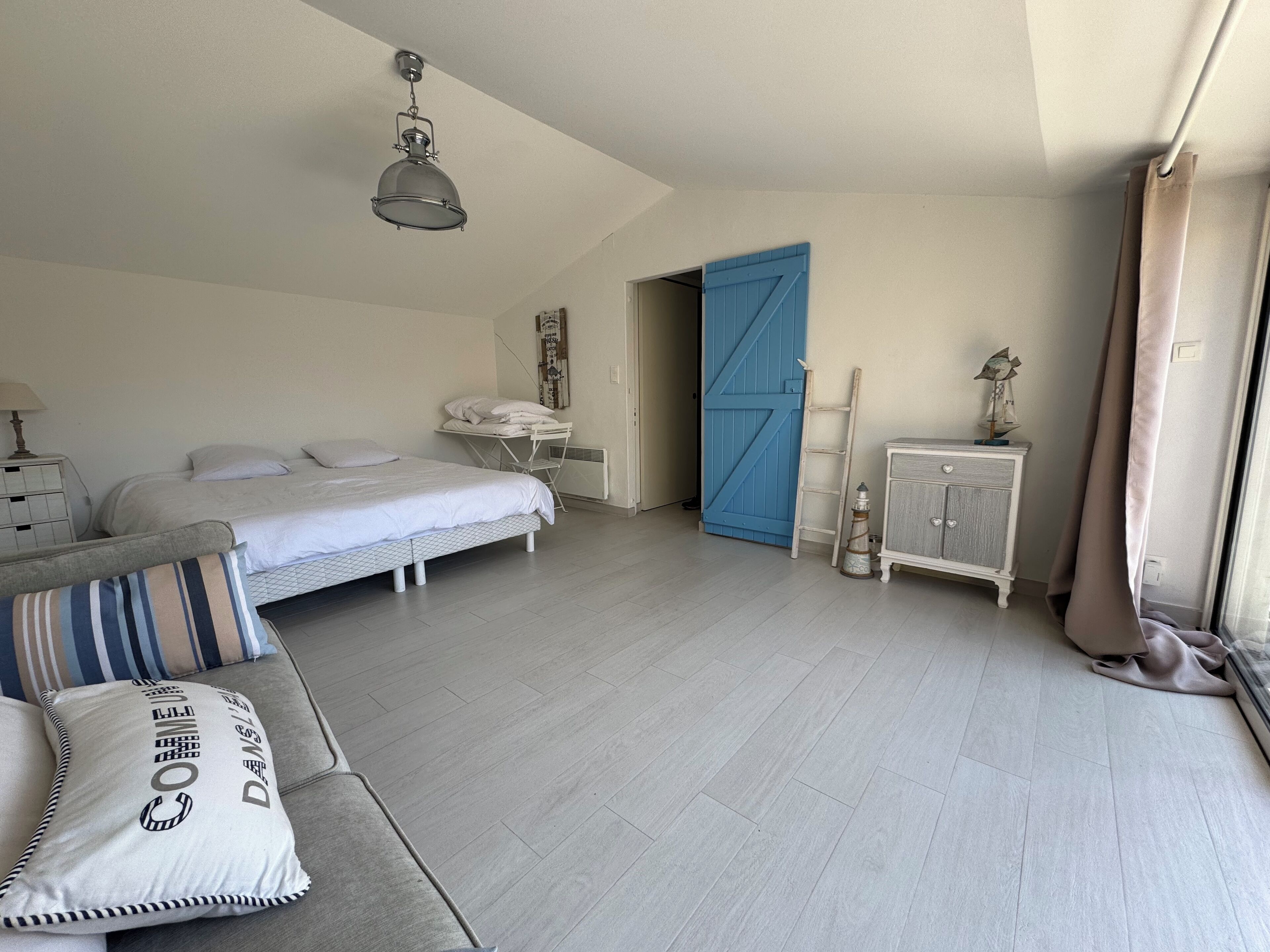 Un dormitorio con cama, cómoda, armario, una puerta azul y una lámpara colgante.