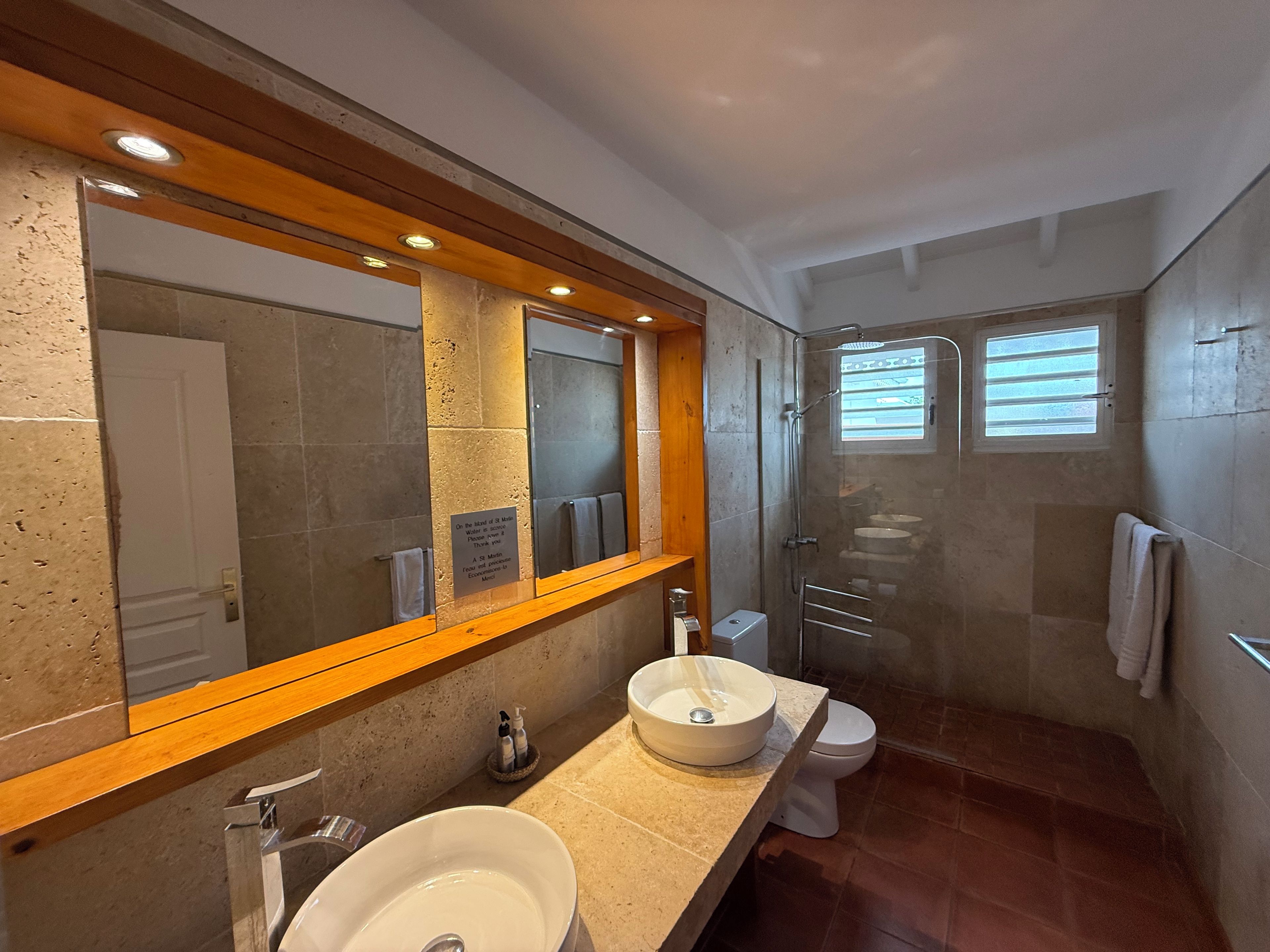 A modern bathroom with a large mirror, two sinks, a shower, and a toilet.