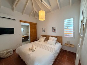 A bedroom with a bed, a bedside table, a hanging light fixture, a window with blinds, and a wall-mounted air conditioning unit. - VILLA SUZANNA in the heart of Orient Bay. Beach and restaurants: 3–4 minutes on foot (baie orientale)