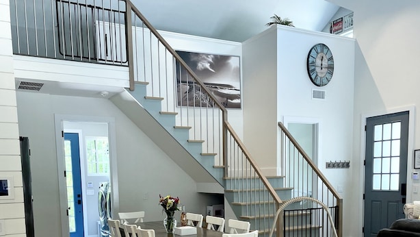 Eine geräumige Küche mit einer zentralen Kochinsel, ein Essbereich mit weißen Stühlen, eine Treppe mit Geländer und eine große Uhr an der Wand.