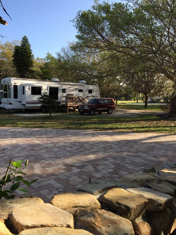 A recreational vehicle (RV) parked on a paved driveway with a stone border, a wooden deck, and a Jeep SUV nearby. - Water View, 100 Fishing Dock! Teton 40 Ft. Trailer W/3 Slide Outs. (Palmetto)