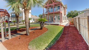 A two-story house with a balcony, surrounded by a wooden fence, palm trees, and a well-maintained lawn.