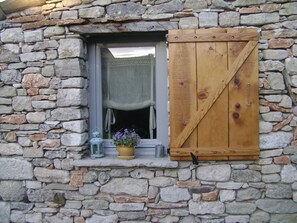 Eine Steinsäule mit einem Fenster, das eine Holzklappe und ein kleines Regal mit einer Laterne und einem Blumentopf zeigt.