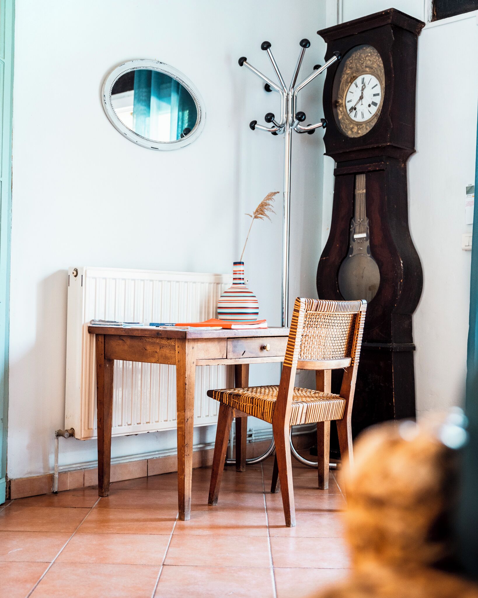 Une pièce comprenant une table en bois, deux chaises en osier, un vase à rayures, un miroir rond et une grande horloge en bois.