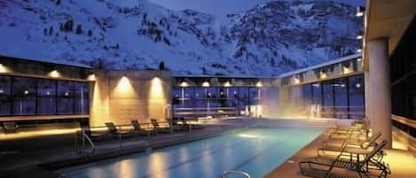 A swimming pool with lounge chairs, surrounded by mountains and a building with large windows.
