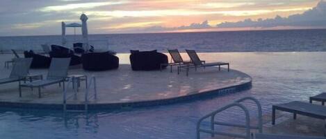 A poolside area with lounge chairs and a small stage, overlooking the sea during sunset.