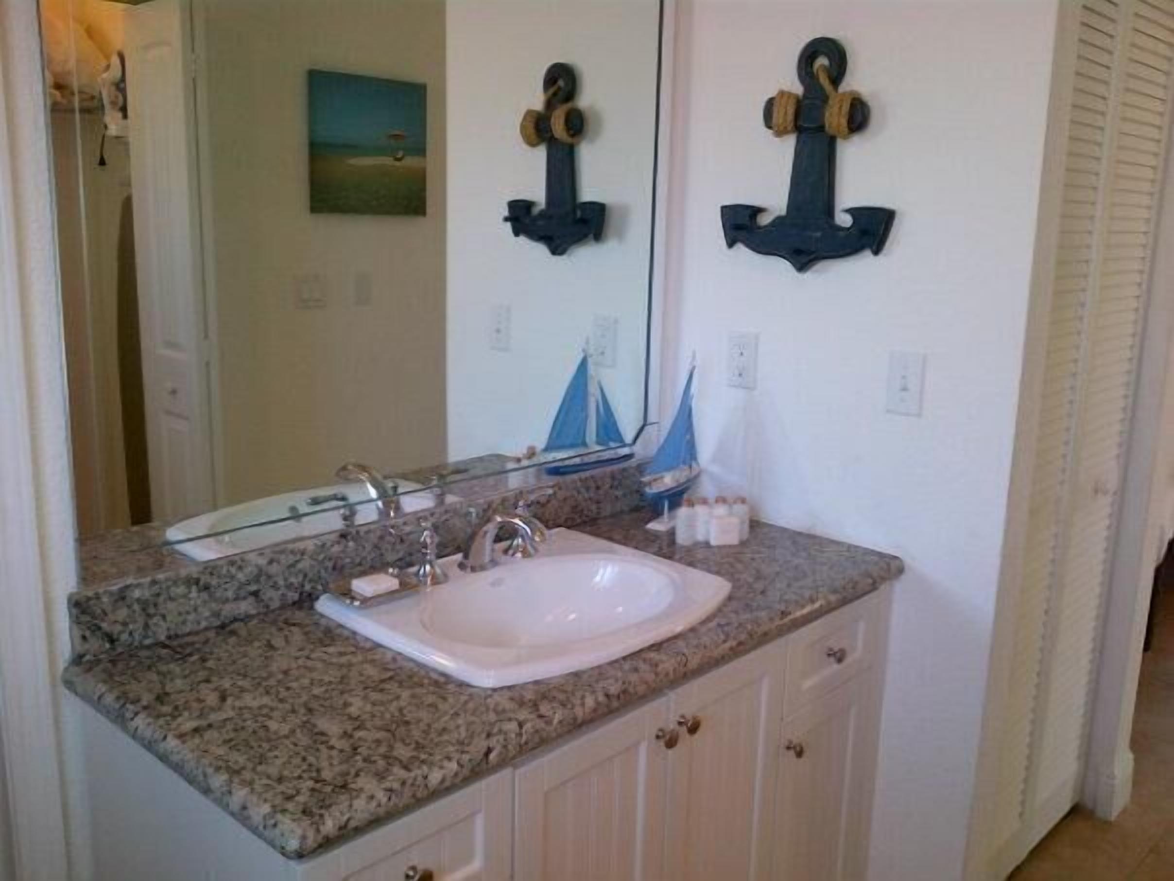 Un baño con encimera de granito, un lavabo blanco y un espejo. Hay dos toalleros de pared y un pequeño barco decorativo.