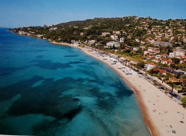 An aerial view of a coastal town with a beach, clear blue waters, and buildings on a hillside.