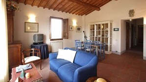 Una sala de estar acogedora con un sofá azul, techo de madera, comedor con sillas y una mesa, y un televisor sobre una mesita auxiliar.