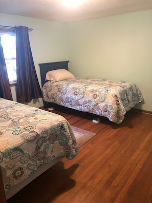 Quarto com duas camas de solteiro, piso de madeira e janela com cortinas.