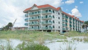 A multi-story hotel with balconies, located near a beach with sandy terrain and sparse vegetation.