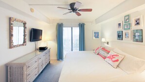 A hotel room with a bed, a dresser, a television, a ceiling fan, and a balcony with curtains.