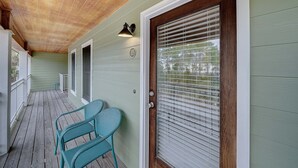 Un porche cubierto con piso de madera, pared exterior verde y una puerta con ventanal.