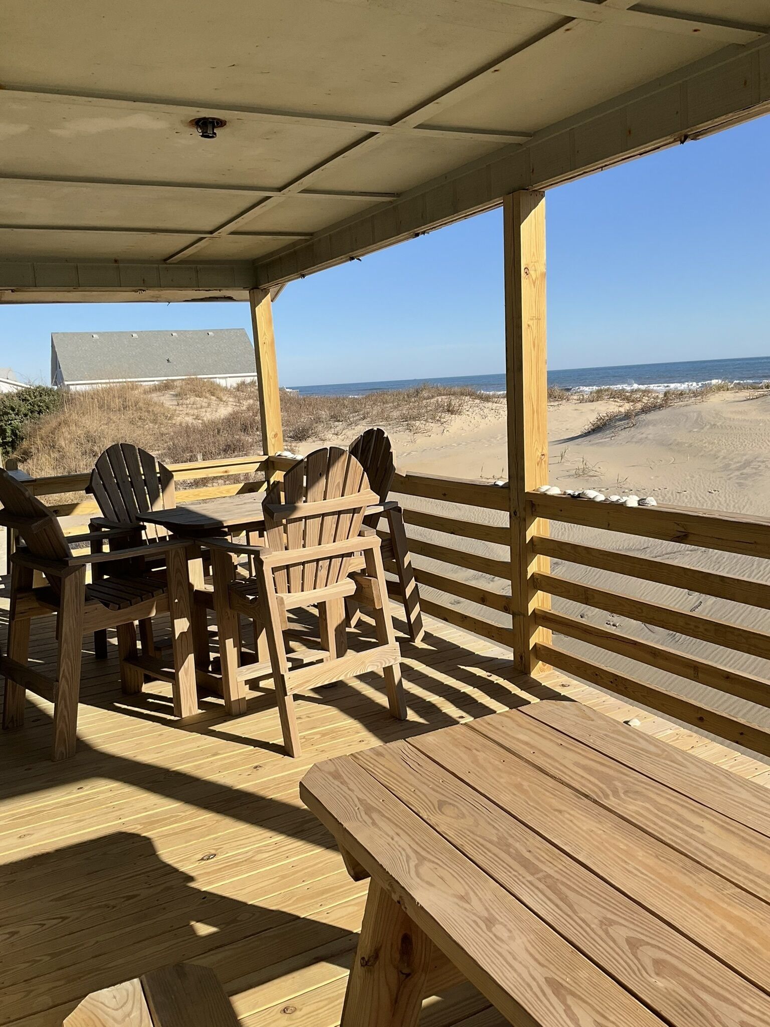 Une terrasse en bois avec une table et des chaises, donnant sur une plage bordée de dunes de sable et sous un ciel dégagé.