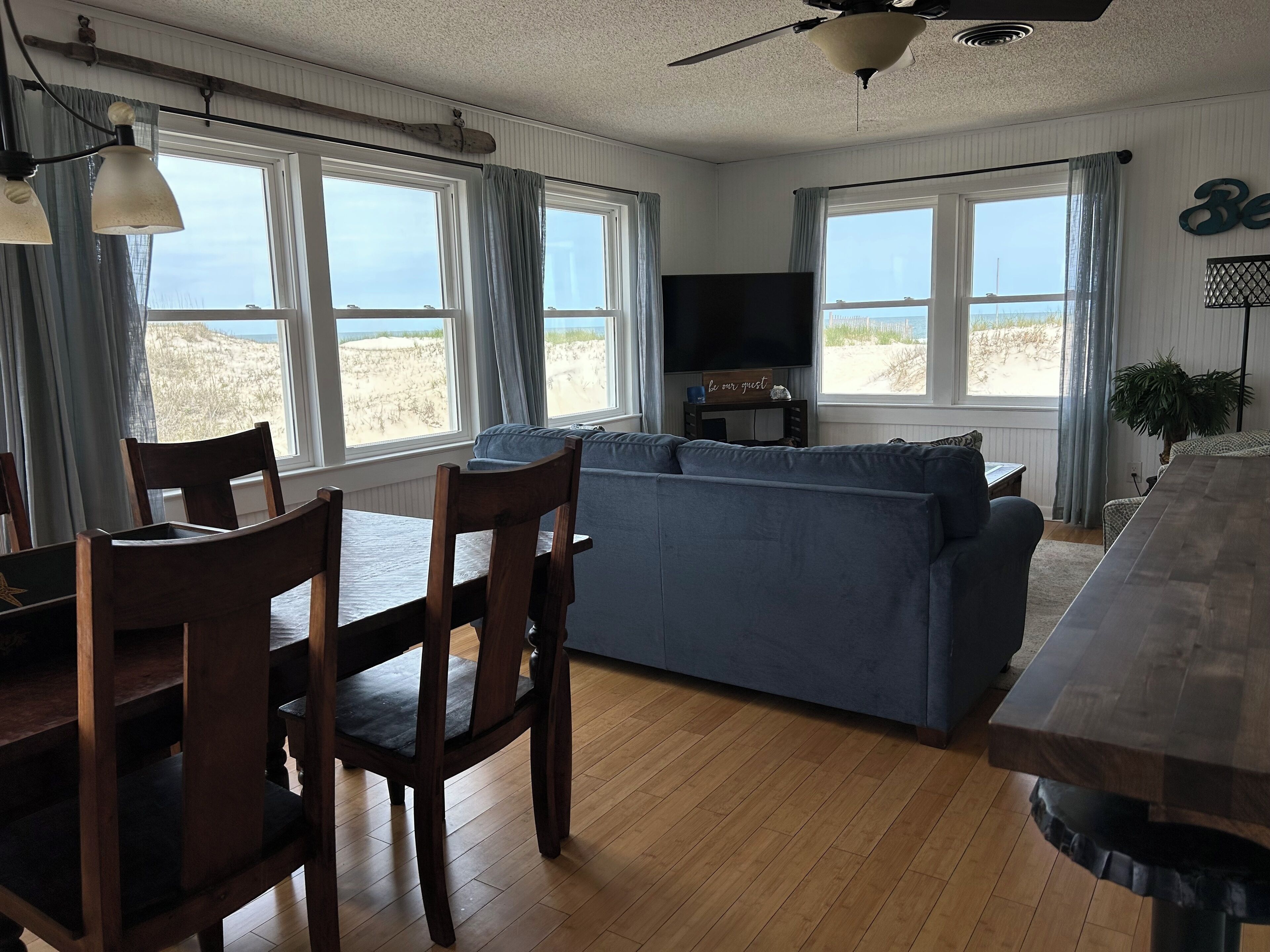 A cozy living room with a blue sofa, wooden dining table, and chairs. There are large windows with curtains, a ceiling fan, and a flat-screen TV on a stand.