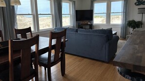 A cozy living room with a blue sofa, wooden dining table, and chairs. There are large windows with curtains, a ceiling fan, and a flat-screen TV on a stand. - SEA GLASS OCEANFRONT COTTAGE.  (Nags Head)