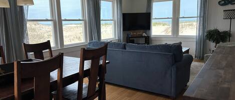 A cozy living room with a blue sofa, wooden dining table, and chairs. There are large windows with curtains, a ceiling fan, and a flat-screen TV on a stand.