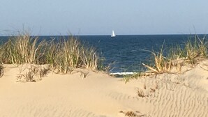 A sandy beach with tall grasses and a sailboat in the distance. - SEA GLASS OCEANFRONT COTTAGE.  (Nags Head)