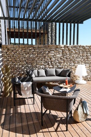 A sunlit outdoor seating area with a stone wall backdrop, a cushioned sofa, wicker chairs, and a table with drinks.