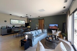 A modern living room with a sofa, coffee table, and TV. There's a dining area with chairs and a table. The room has a large window and a ceiling fan.