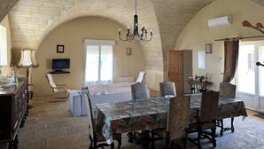 Un comedor con una mesa preparada para cuatro personas, rodeada de sillas tapizadas. Un candelabro cuelga del techo y un televisor está montado en la pared.