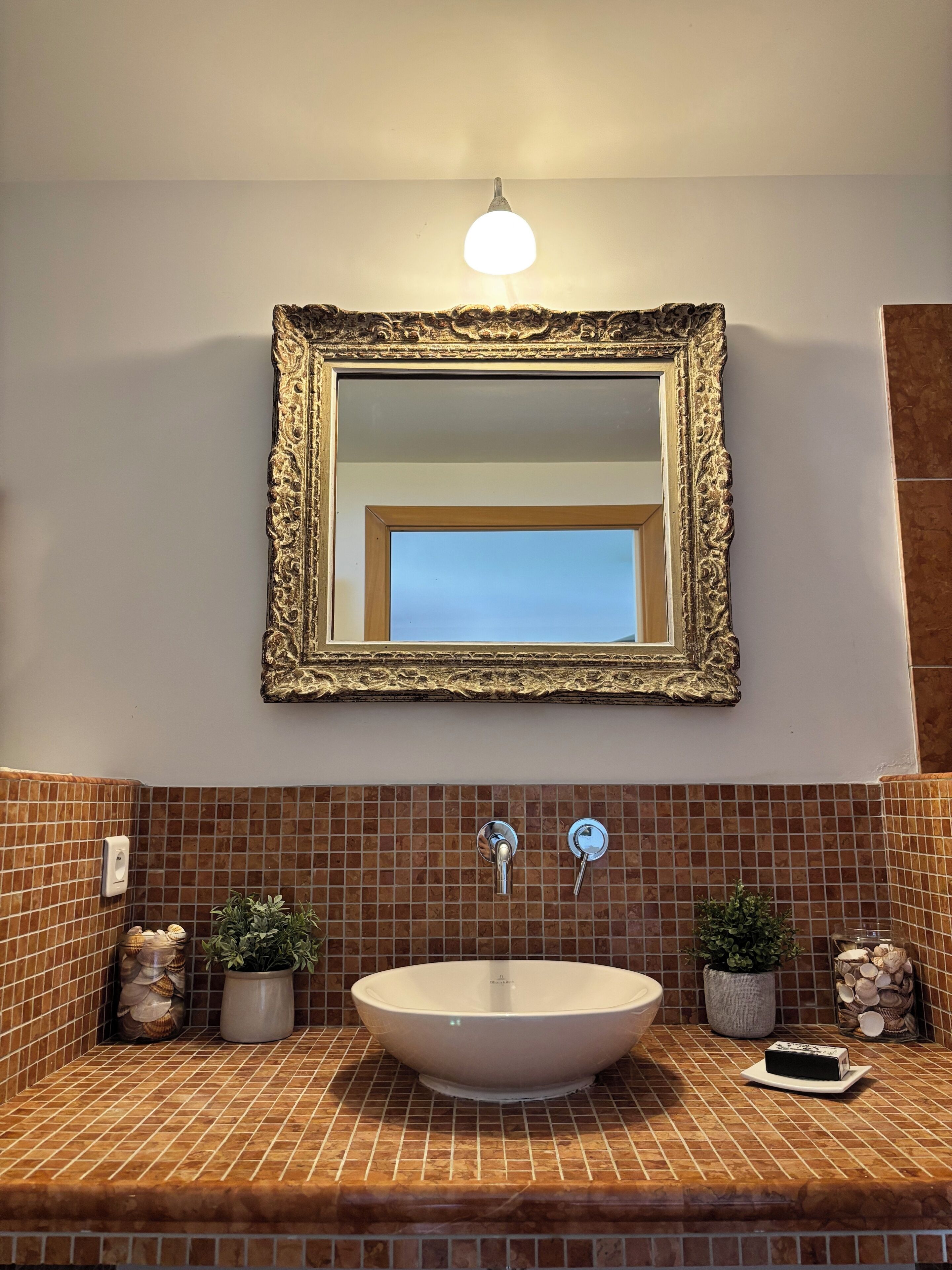 Une salle de bain avec un grand lavabo ovale, un miroir encadré d’or et deux plantes en pot.