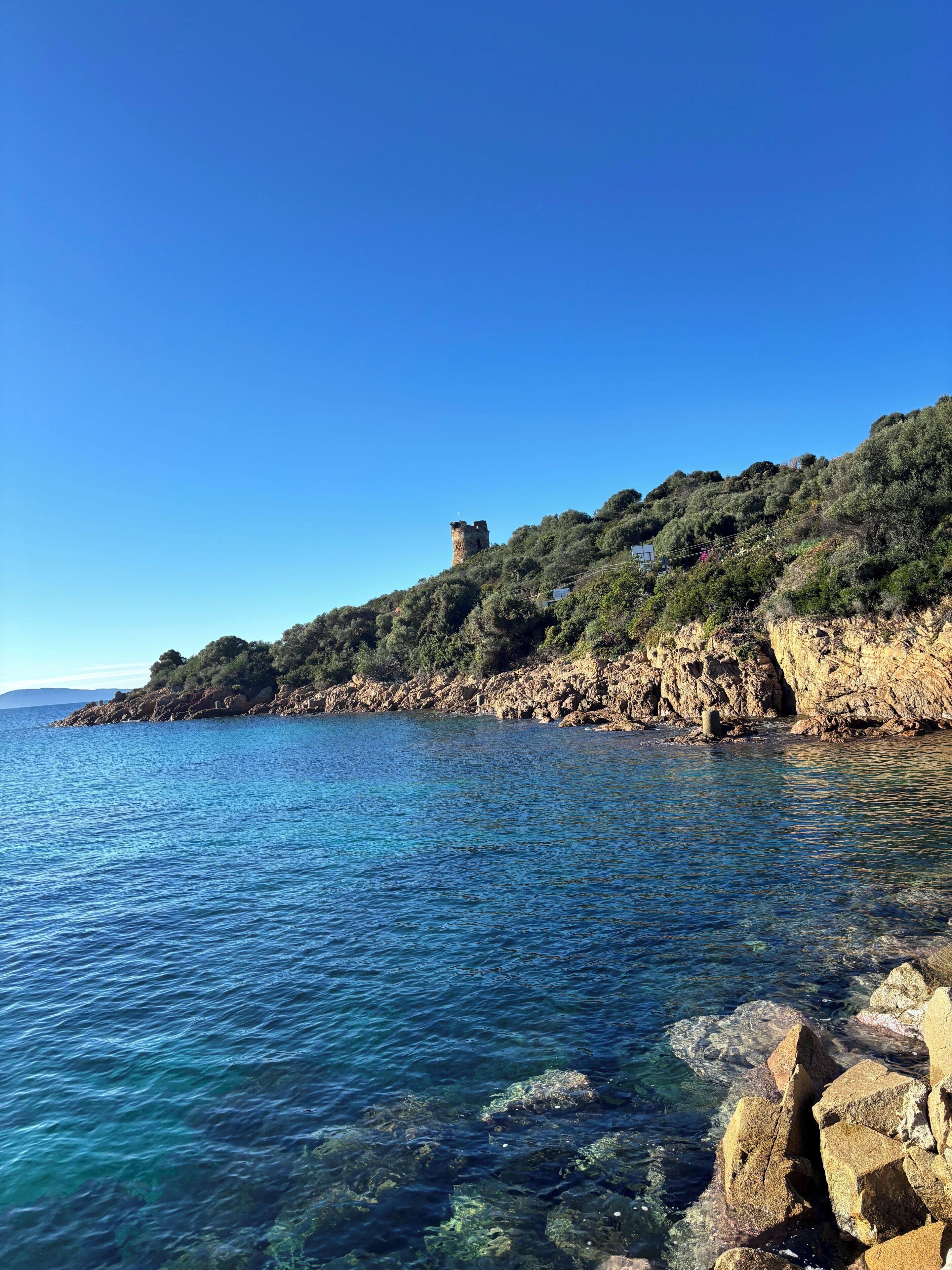 Paesaggio costiero con un litorale roccioso, acqua blu cristallina e una torre su una collina.