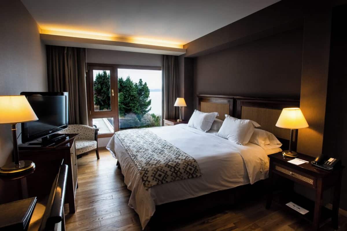 A hotel room with a large bed, two bedside lamps, a TV, a desk with a lamp, and a view of greenery outside.