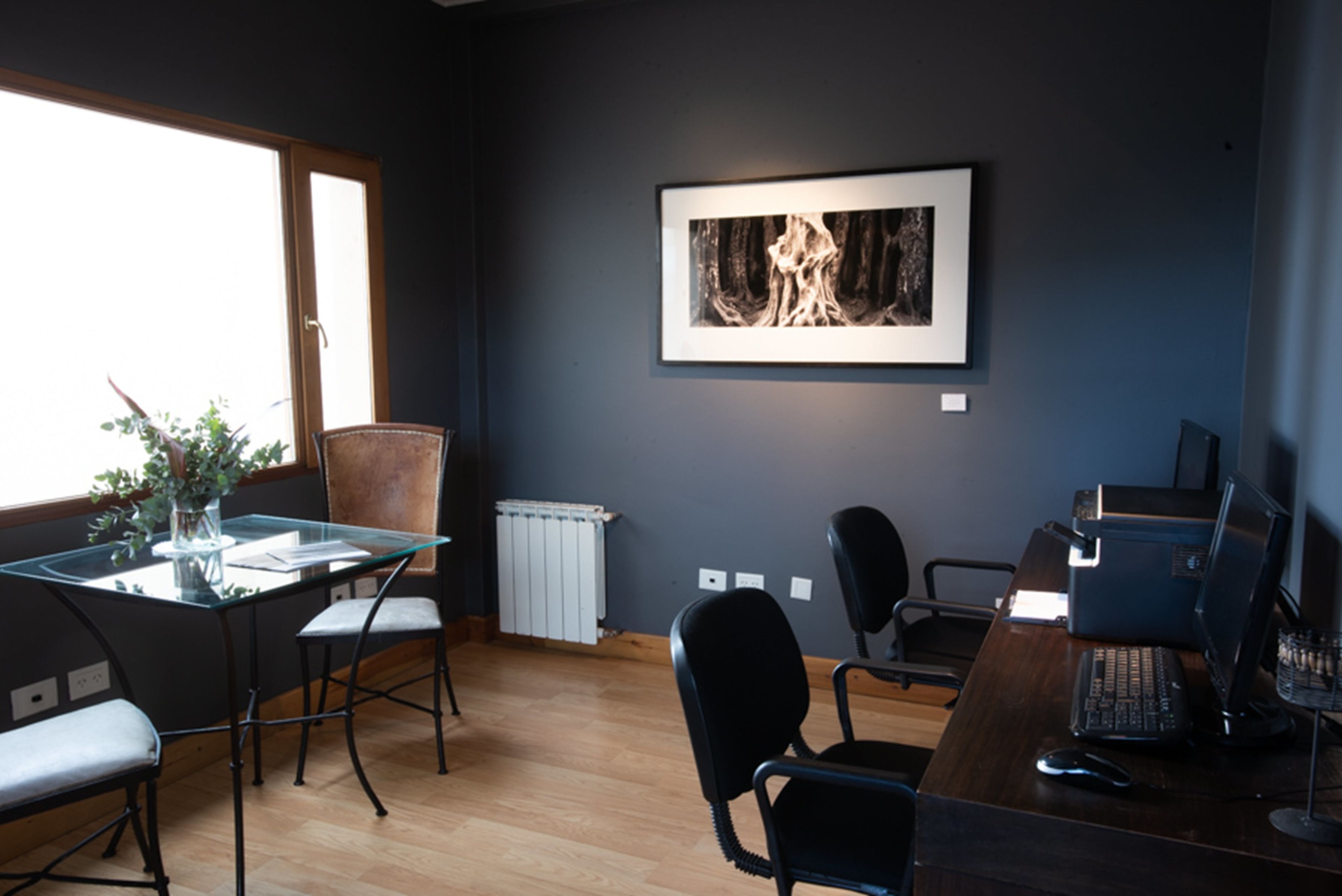A modern office with a glass table, black chairs, a computer desk, and a framed picture on the wall.