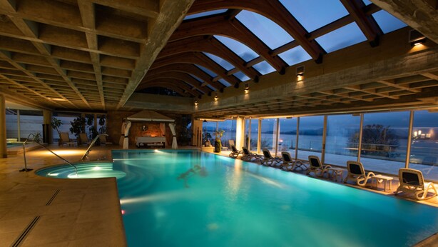 Una piscina interior amb vistes a l'exterior, sostre de fusta i seients de relaxació al costat de la piscina.