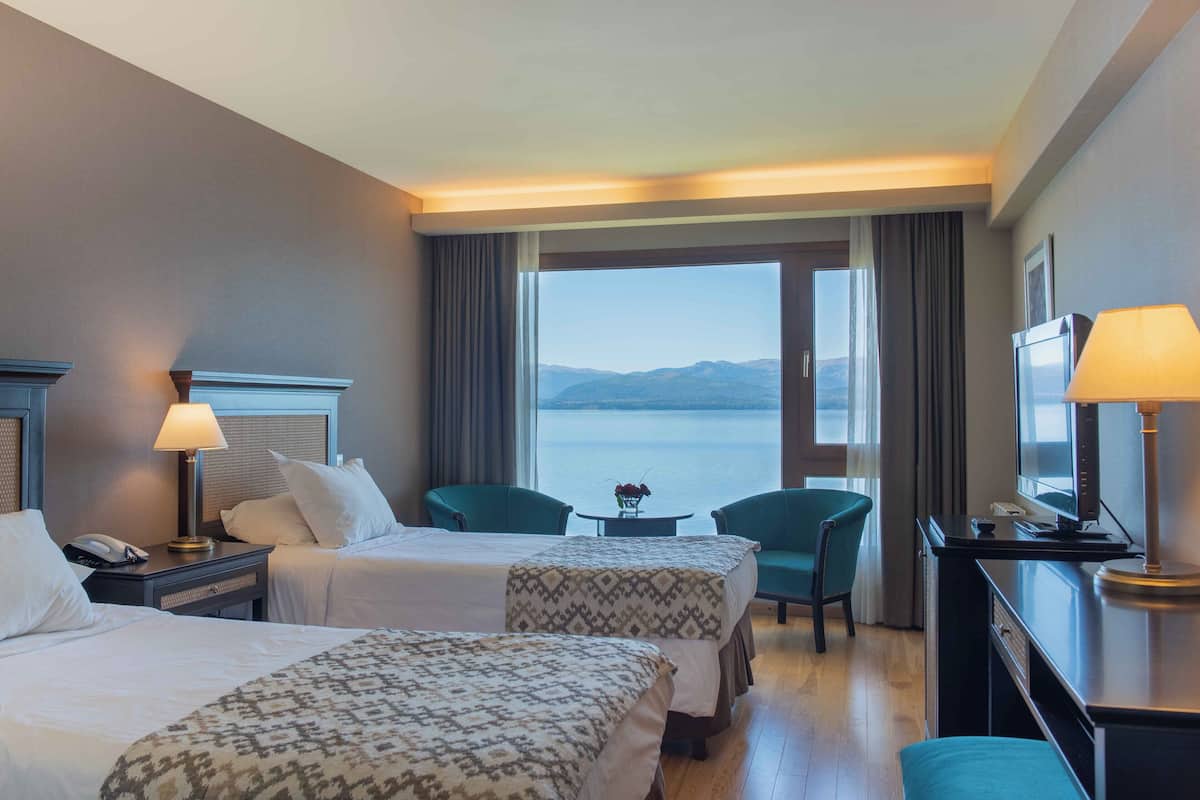 A hotel room with two beds, a desk with a lamp, a chair, and a view of the water.
