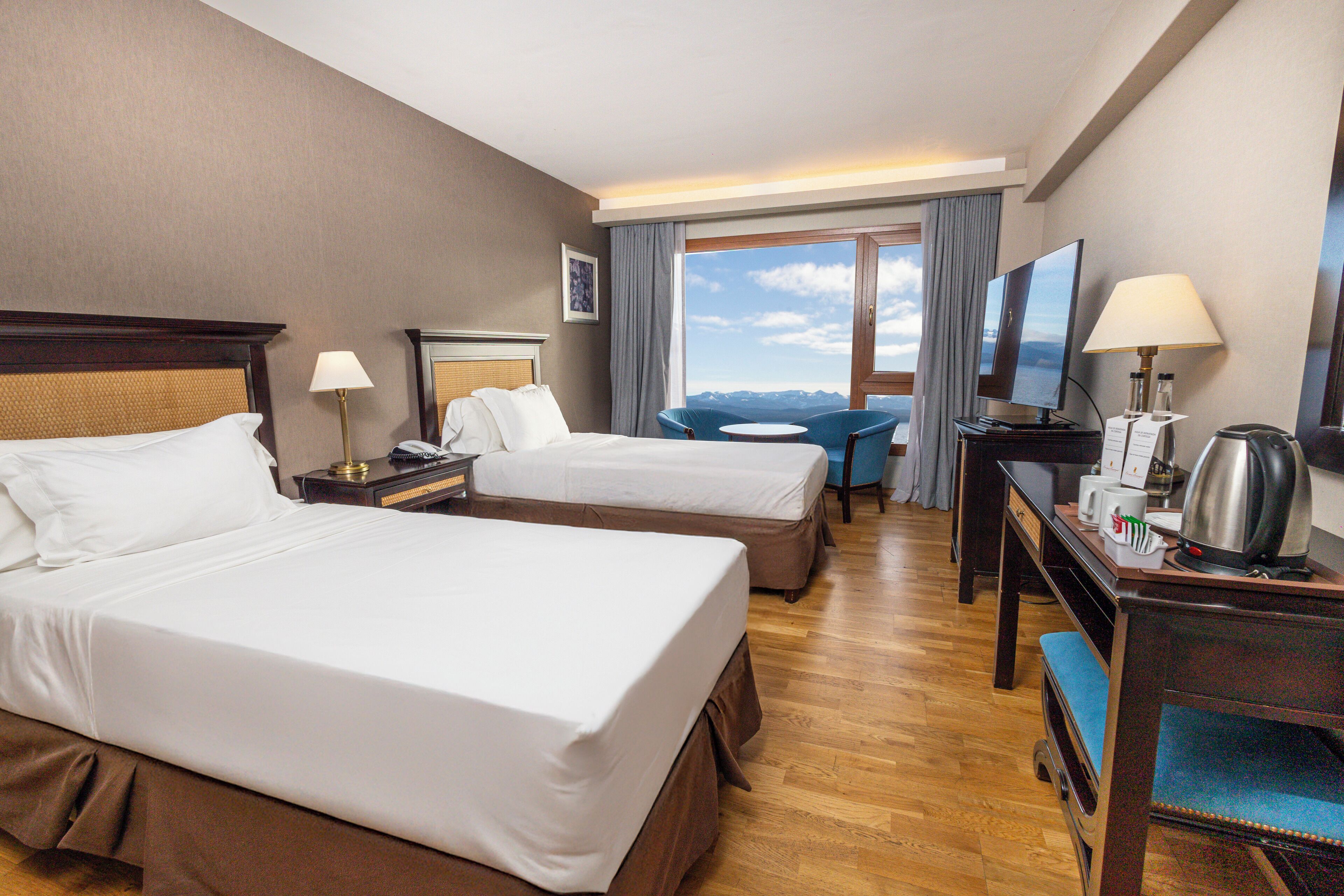 Quarto de hotel com duas camas, uma escrivaninha com cadeira, televisão e janela com vista.