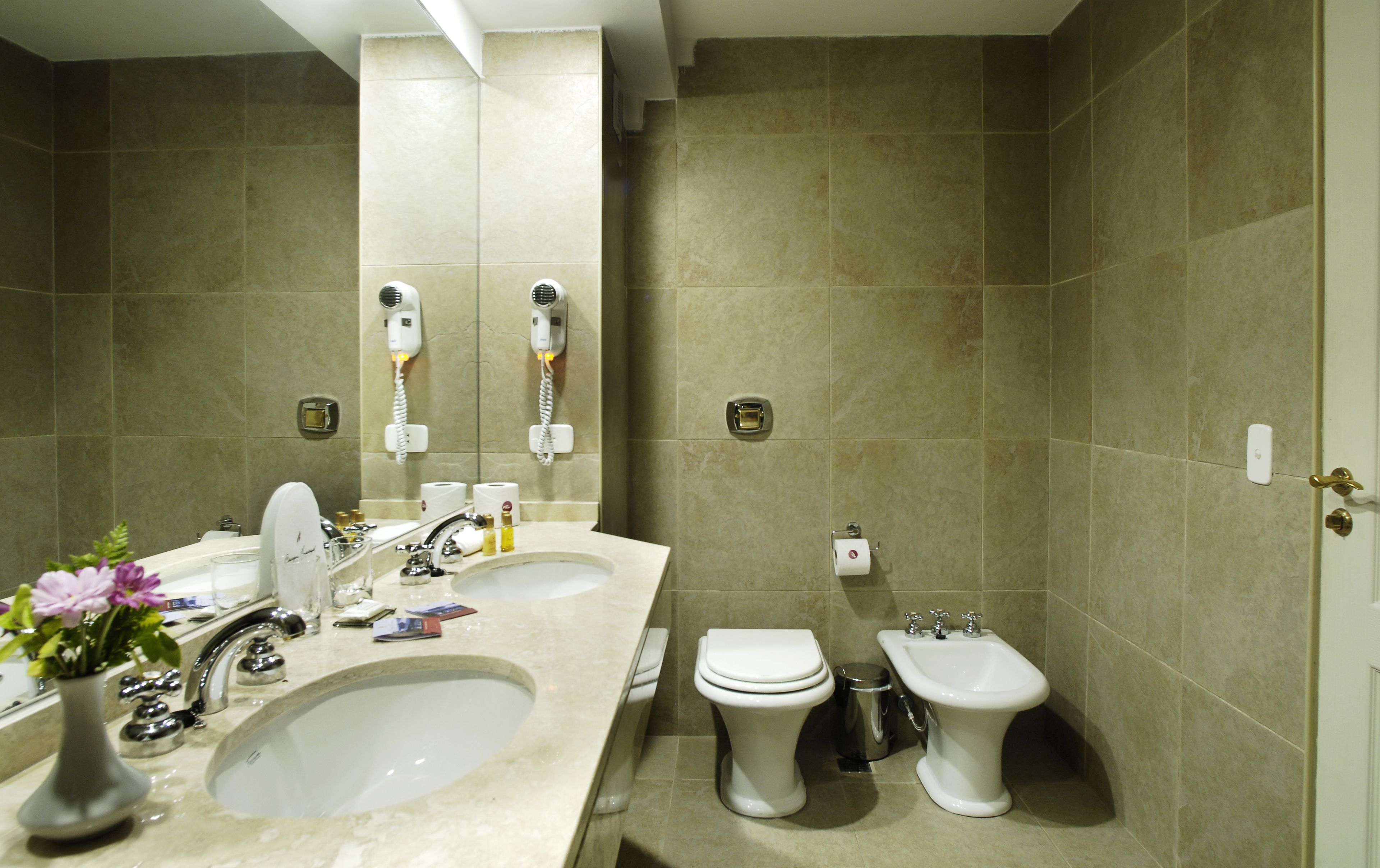 Un baño con dos lavamanos, un inodoro y un bidé. Hay secadores de pelo fijados a la pared.
