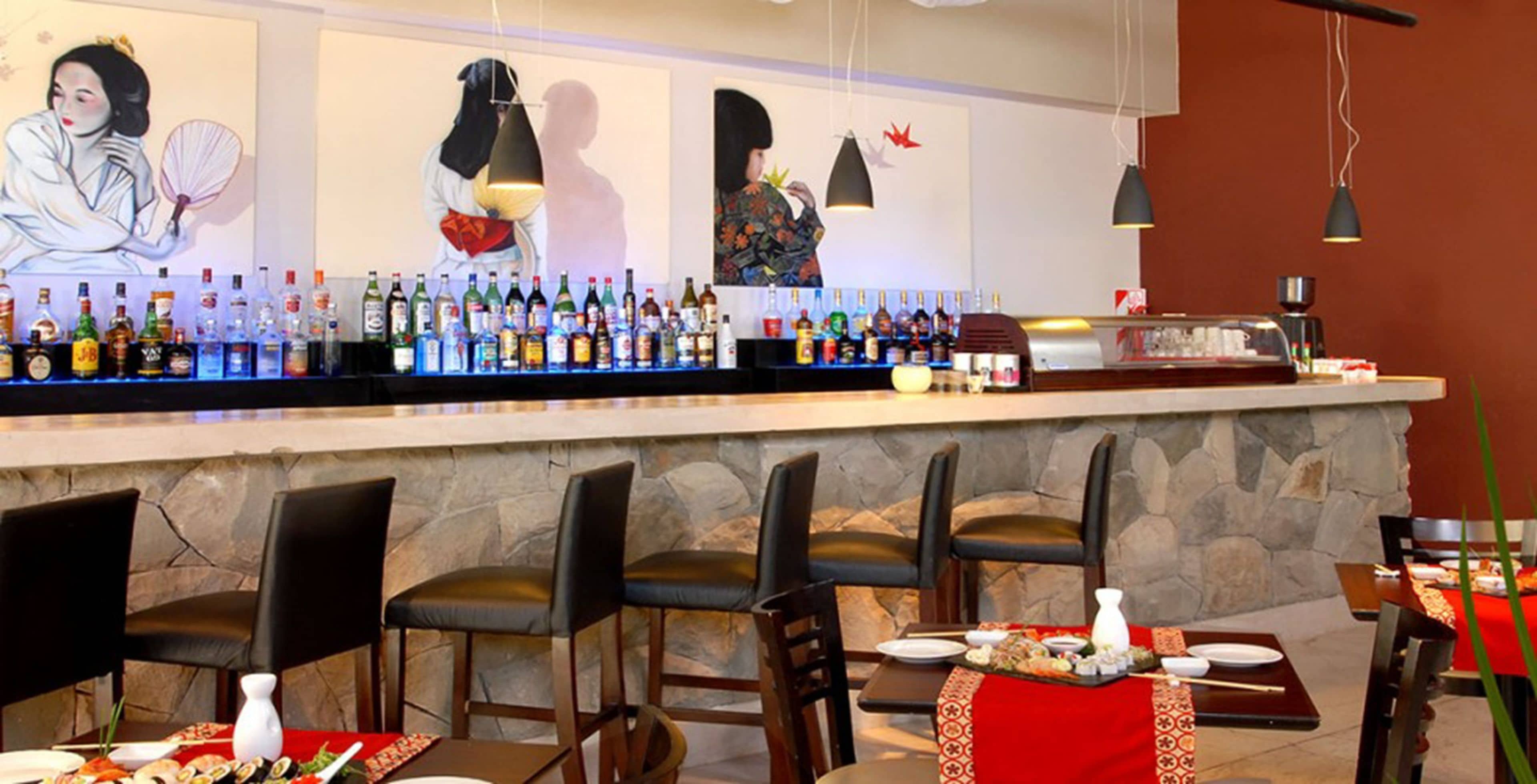 Zona de bar con pared de piedra, diversas botellas de licor y diferentes opciones de asientos, incluyendo taburetes y mesas.
