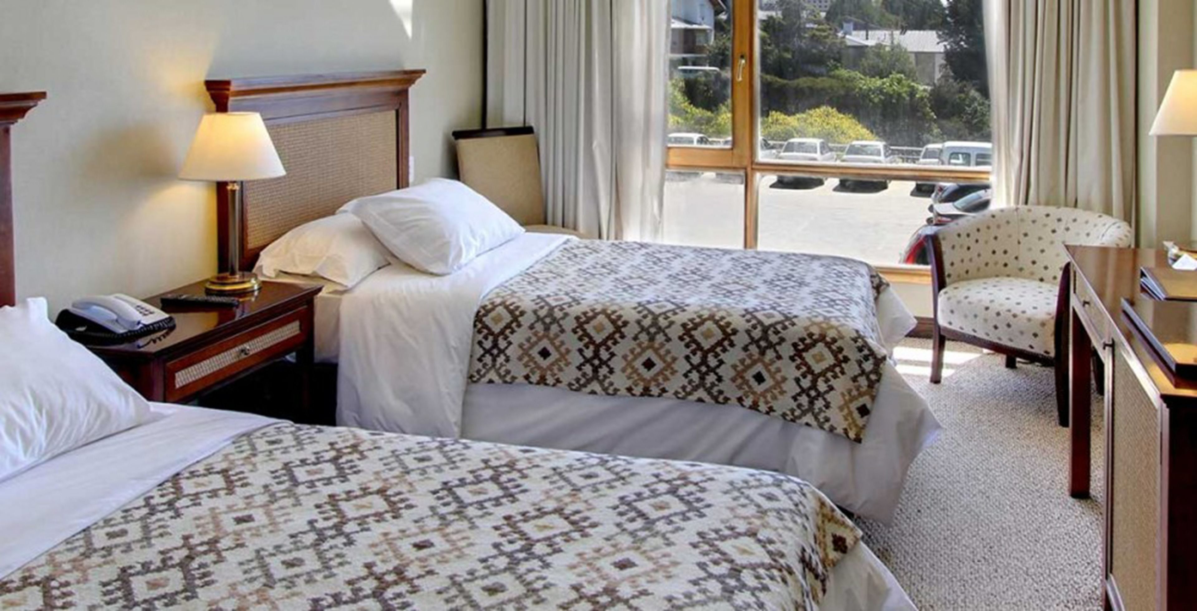A hotel room with two beds, a desk, a chair, and a window with a view of parked cars and greenery outside.