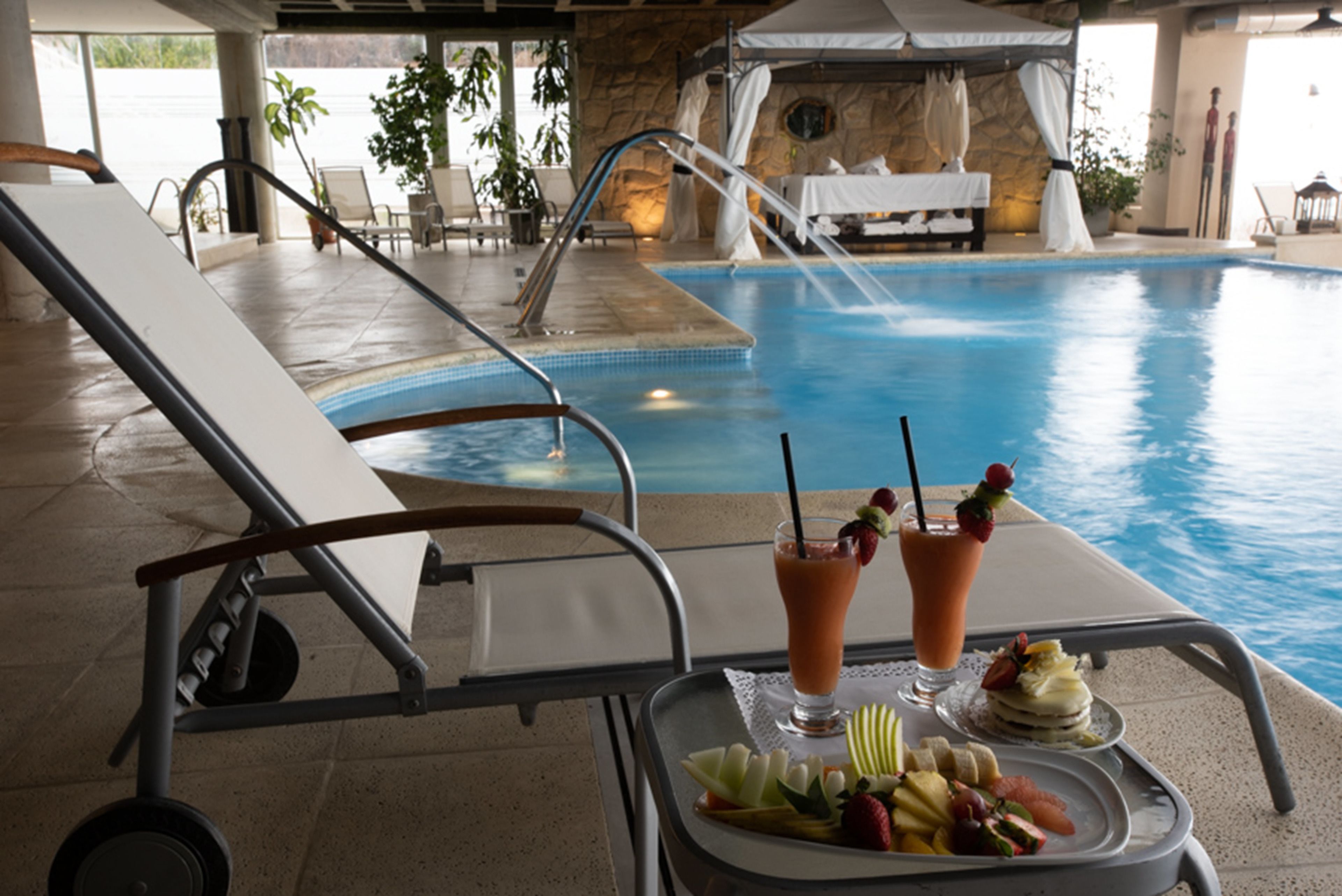Uma área à beira da piscina com espreguiçadeiras, uma bandeja com frutas e bebidas, e vista para a piscina.